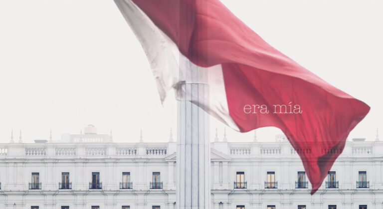 Imagen de tonalidad blanquecina, horizontal. En la parte superior el cielo blanco ocupa dos tercios de la imagen, al fondo se muestra la última planta del “Palacio de La Moneda”. Al centro un asta gruesa blanca sostiene la bandera chilena movida por el viento, solo se muestra la zona blanca y roja en la que está escrito “era mía”.