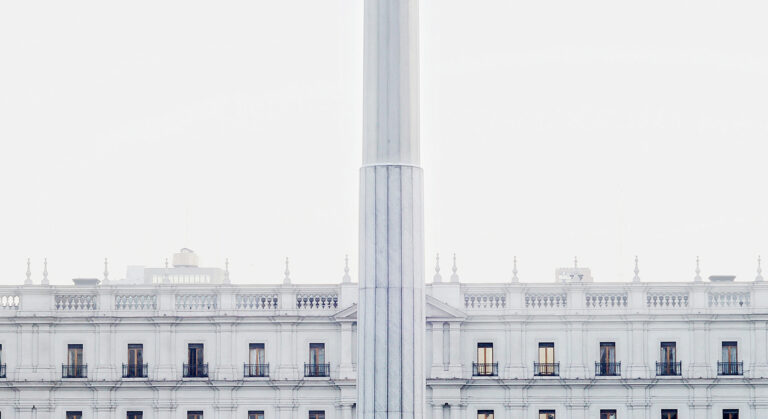 Imagen de tonalidad blanquecina, horizontal. En la parte superior el cielo blanco, al fondo se muestra la planta superior del edificio “Palacio de La Moneda”. Al centro una asta gruesa blanca.