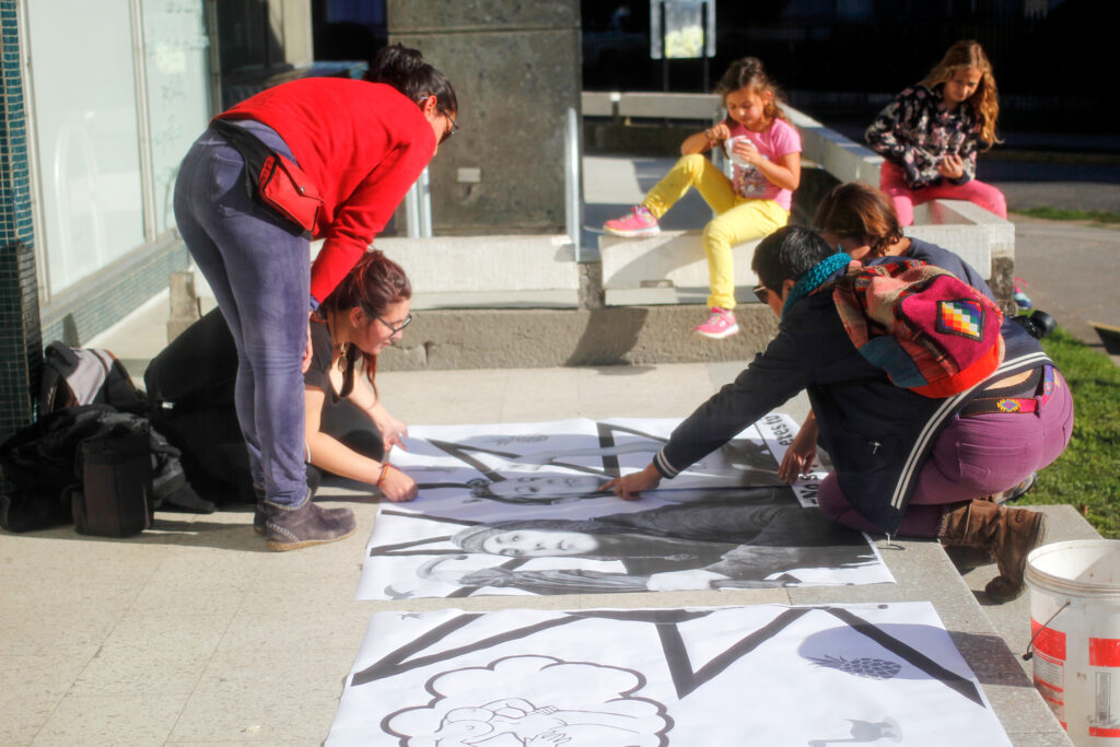 Grupo de cuatros mujeres juntan grandes pliegos de papel impresos en blanco y negro sobre baldosas de las afueras de la Pinacoteca de la Universidad de Concepción, en un día soleado.