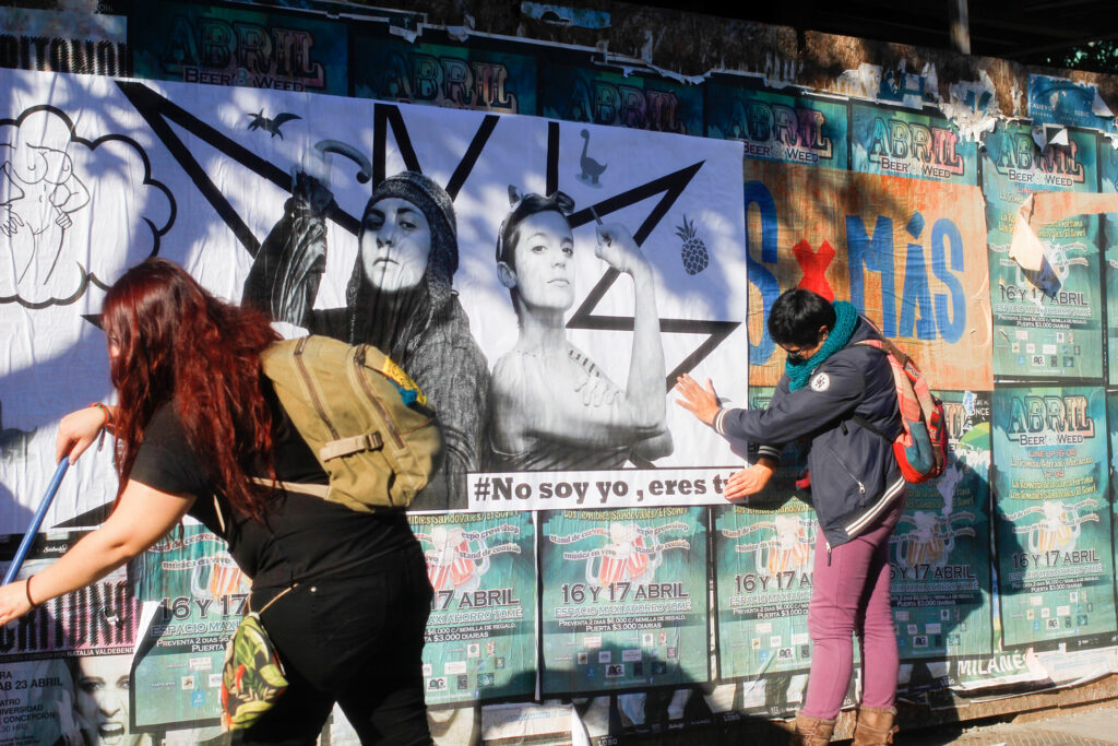 Dos mujeres pegan una gigantografía en un muro lleno de afiches verdes, donde aparecen dos mujeres en posición de autodefensa, una vestida con ropa de invierno con un paraguas en las manos y la otra con polera manga corta y una llave empuñada. A la izquierda una mujer joven está con un escobillón en la mano y otra estirando el papel sobre el muro, donde dice #nosoyyo,erestú