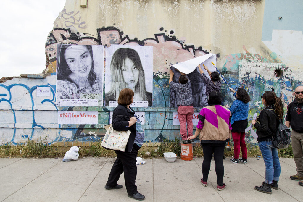 Grupo de personas pegando gigantografias de mujeres jóvenes asesinadas en el año 2016: Alison Calderon Hidalgo 17 años - Santiago de Chile y Yenery Carrasco González 18 años. Dos personas están pegando una tercera imágen. Se ve escrito #niunamenos. En primer plano una mujer mayor mira lo que se está haciendo.