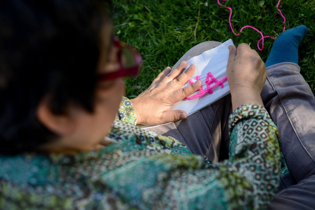 Detalle del bordado de una mujer que está sin zapatos sobre el pasto. La fotografía es tomada desde arriba y tras la mujer, enfocando las manos que sostienen el bordado de color fucsia.