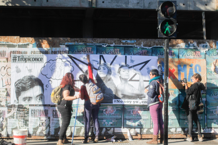 Grupo de mujeres pegando una gran imagen de papel en blanco y negro en una zona urbana. En ella aparece un joven imaginado a una mujer desnuda dibujada como historieta y dos mujeres jóvenes: la primera en posición de defensa con un paraguas, viste ropa de invierno y la otra, toma como arma de defensa una llave entre los dedos con su mano empuñada, viste ropa de verano.