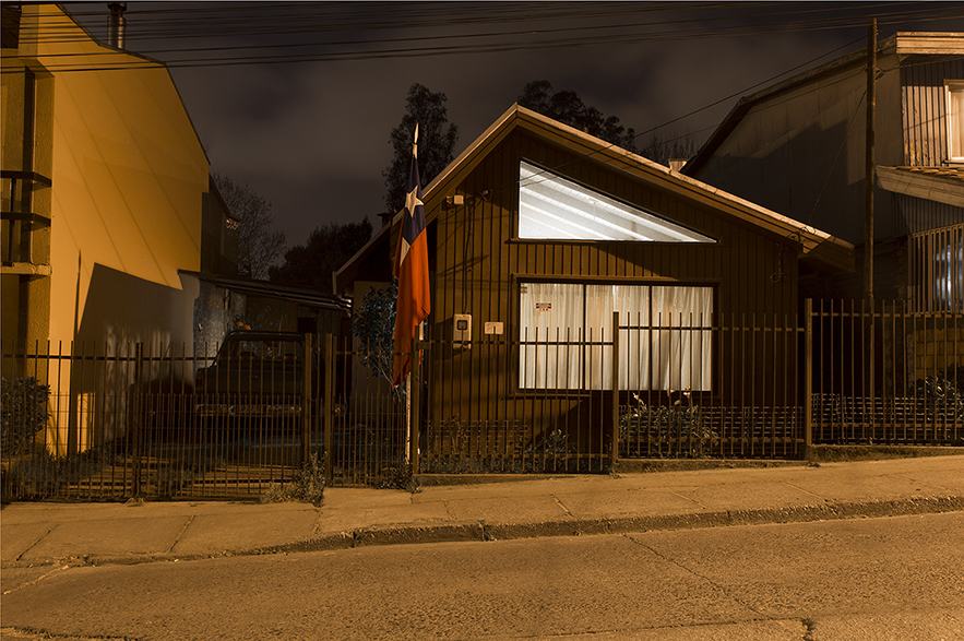 Esta imagen horizontal muestra una casa en un entorno urbano, tomada durante la noche. La escena está iluminada por luces artificiales y la casa presenta una bandera chilena en el exterior. La casa tiene un diseño sencillo y funcional, con una cerca que delimita la propiedad.