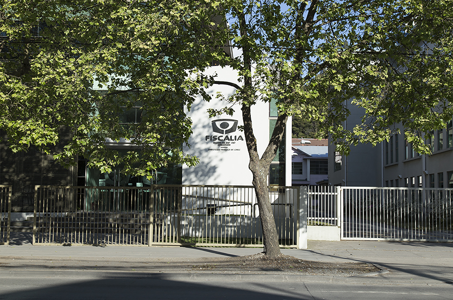 Fotografía de la fiscalía de la ciudad de Concepción. Imagen horizontal a color, al fondo un edificio blanco lleva escrito: Fiscalía, con reja blanca que resguarda toda la fachada. Al lado izquierdo está la entrada de peatones y al lado derecho la de vehículos. En la parte inferior la vereda y casi al centro un árbol delgado con muchas hojas verdes.