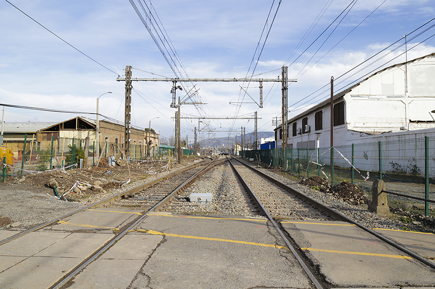 Fotografía a color de zona urbana de la ciudad de San Fernando. Imagen horizontal que muestra un camino férreo. En la mitad superior hay un cielo celeste con nubes blancas donde se observan tendidos eléctricos y cables del tren que se pierden en una perspectiva profunda. Al centro 2 líneas férreas y en primer plano un camino de cemento.