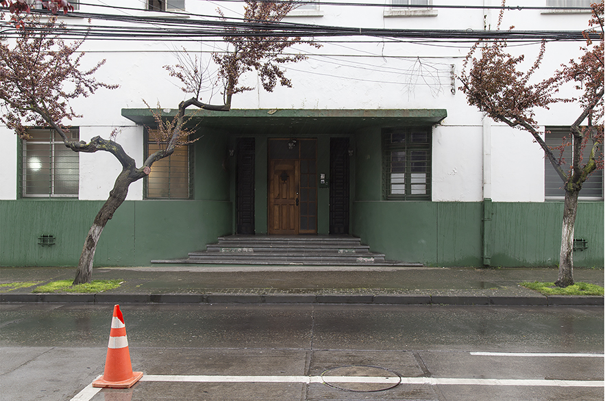 Fotografía horizontal a color. Muestra una comisaría de Carabineros de Chile, de la ciudad de Concepción. Construcción mitad blanca y verde musgo. En el frontis hay cuatro ventanas con rejas y entrada protegida con un alero, sobre la vereda 2 árboles con pocas hojas y sobre ellos gruesos cables eléctricos. En la parte inferior, la calle mojada.