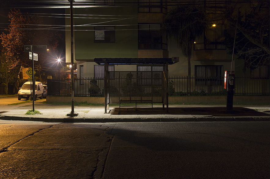 Fotografía de zona urbana con poca iluminación de la ciudad de Concepción. Imagen horizontal a color. Al fondo un edificio habitacional blanco protegido por una reja. En la vereda, que tiene muy poca iluminación, hay un paradero en penumbra.