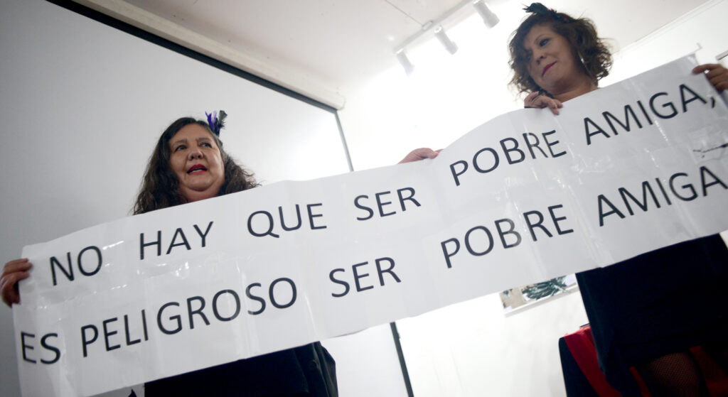 Dos mujeres vestidas de negro sostienen un lienzo de papel impreso que dice: “No hay que ser pobre amiga, es peligroso ser pobre amiga”. Son las poetas Bárbara Calderón y Rosy Sáez del colectivo “¿Por qué tan solitas? La fotografía es tomada desde el suelo hacia arriba, al fondo un telón y pared blanca. Lugar: Casa 916 en Concepción.