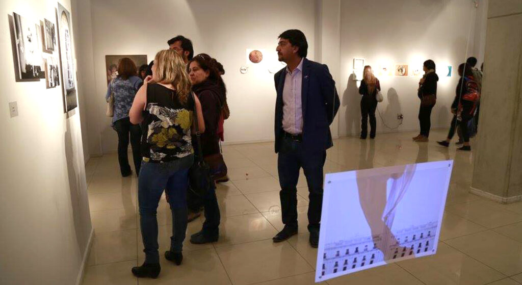 Exposición fotográfica, diversos grupos de personas observan y comentan las imágenes expuestas. En primer plano el video de la obra Marcela Bruna, proyectada sobre papel colgando con hilo transparente.