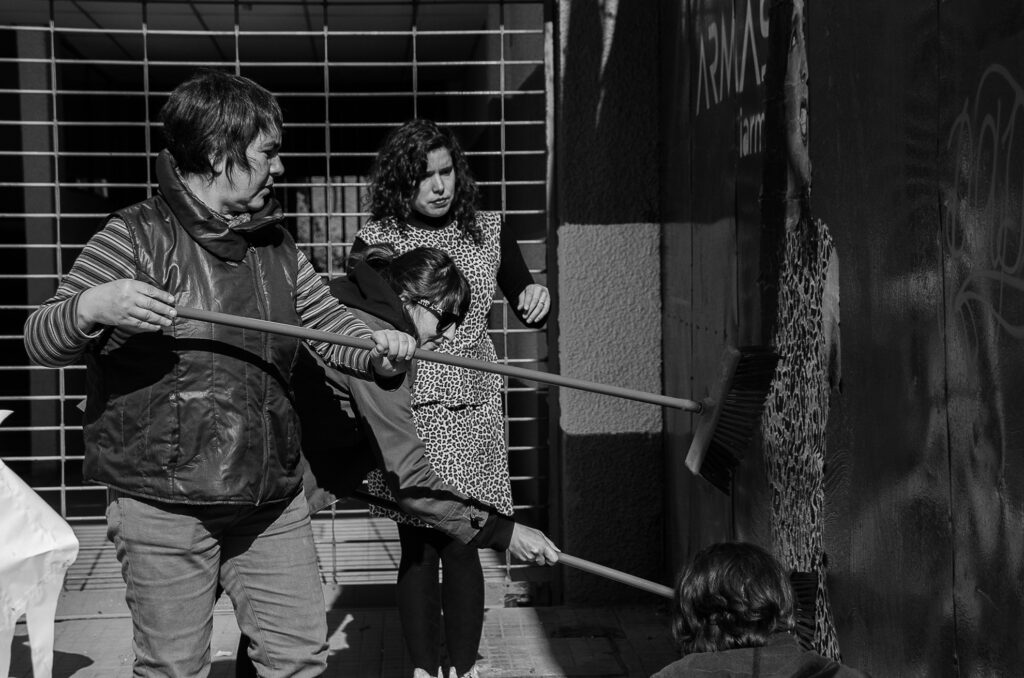 Grupo de personas pegando sobre un muro una imagen fotográfica en gran escala realizada para la accion “El dedo de Jara”. Dos mujeres están de pie pasando sobre la imagen una escoba para reforzar el pegamento sobre la muralla. Al costado derecho se aprecia la imagen que es superior a la escala humana.